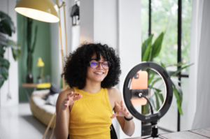 A phone and ring light set up to record a young female influencer in a bright room with windows, plants and modern furniture.