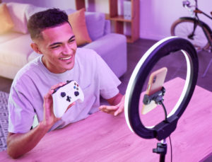 a young man recording a vertical video with a phone and ring light setup. He is holding a gaming controller and is sitting in a modern living room lit with pink LED lights.